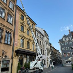 Usługi Podnośnikami Koszowymi Krzysztof Miłoński. - Podnośnik koszowy Ruthmann na tle kamienic Starego Miasta w Warszawie, z numerem telefonu i adresem strony internetowej na wysięgniku.