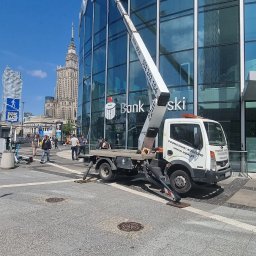 Usługi Podnośnikami Koszowymi Krzysztof Miłoński. - Pracownik w koszu na wysięgniku przy szklanej fasadzie budynku banku w centrum miasta, w tle Pałac Kultury i Nauki.