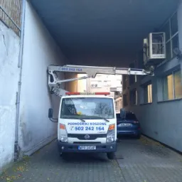 Biały samochód z wysięgnikiem parkuje w wąskim przejeździe między budynkami, platforma z pracownikiem unosi się przy ścianie budynku z oknami.