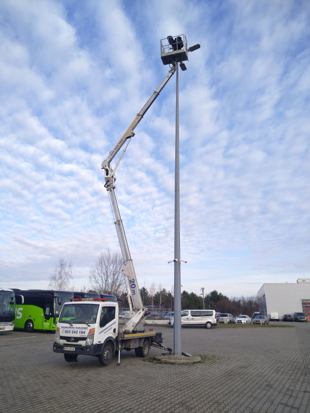 Wysięgnik koszowy na pojeździe ustawiony obok wysokiego słupa oświetleniowego z zamontowanymi reflektorami, na tle pochmurnego nieba. Na pierwszym planie zaparkowane autobusy i busy.
