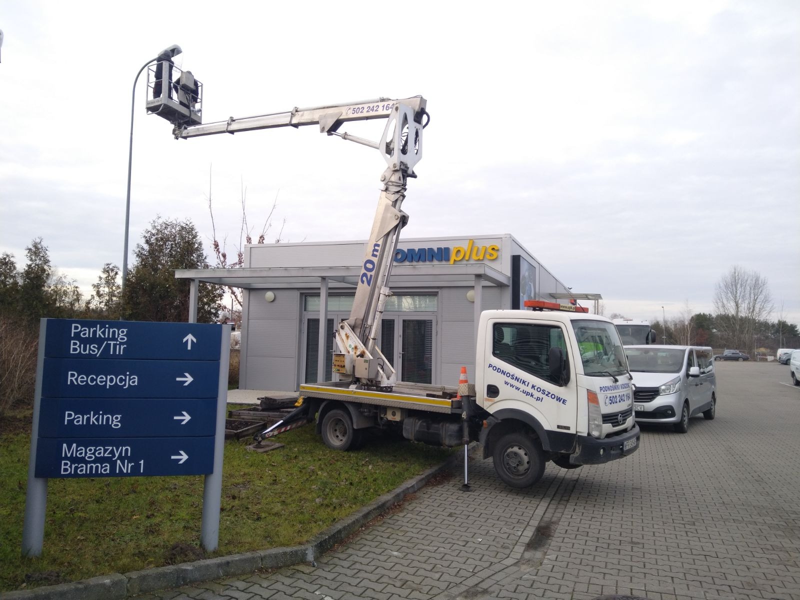 Praca na wysokościach: podnośnik zamontowany na samochodzie na tle budynku Omniplus i tablicy informacyjnej z napisem Parking Bus/Tir.