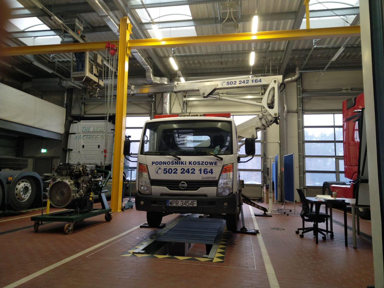 Ciężarówka z zamontowanym ramieniem, zaparkowana nad kanałem w warsztacie, obok silnik na wózku i podnośnik na szynie pod sufitem.