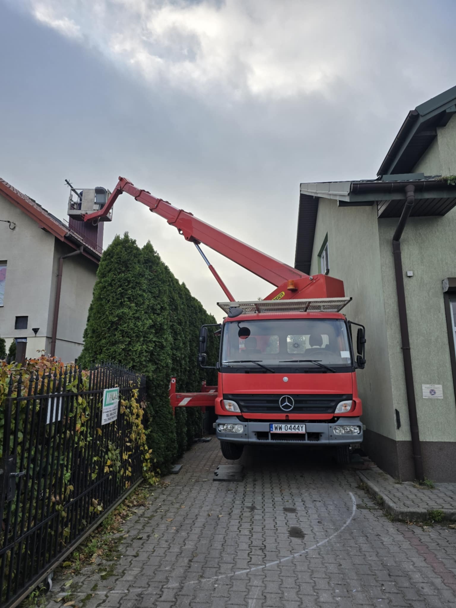 Czerwony pojazd z wysięgnikiem roboczym Mercedes-Benz zaparkowany na brukowanym podjeździe między budynkami, wysięgnik uniesiony w kierunku dachu, tablica rejestracyjna WW 0444Y.