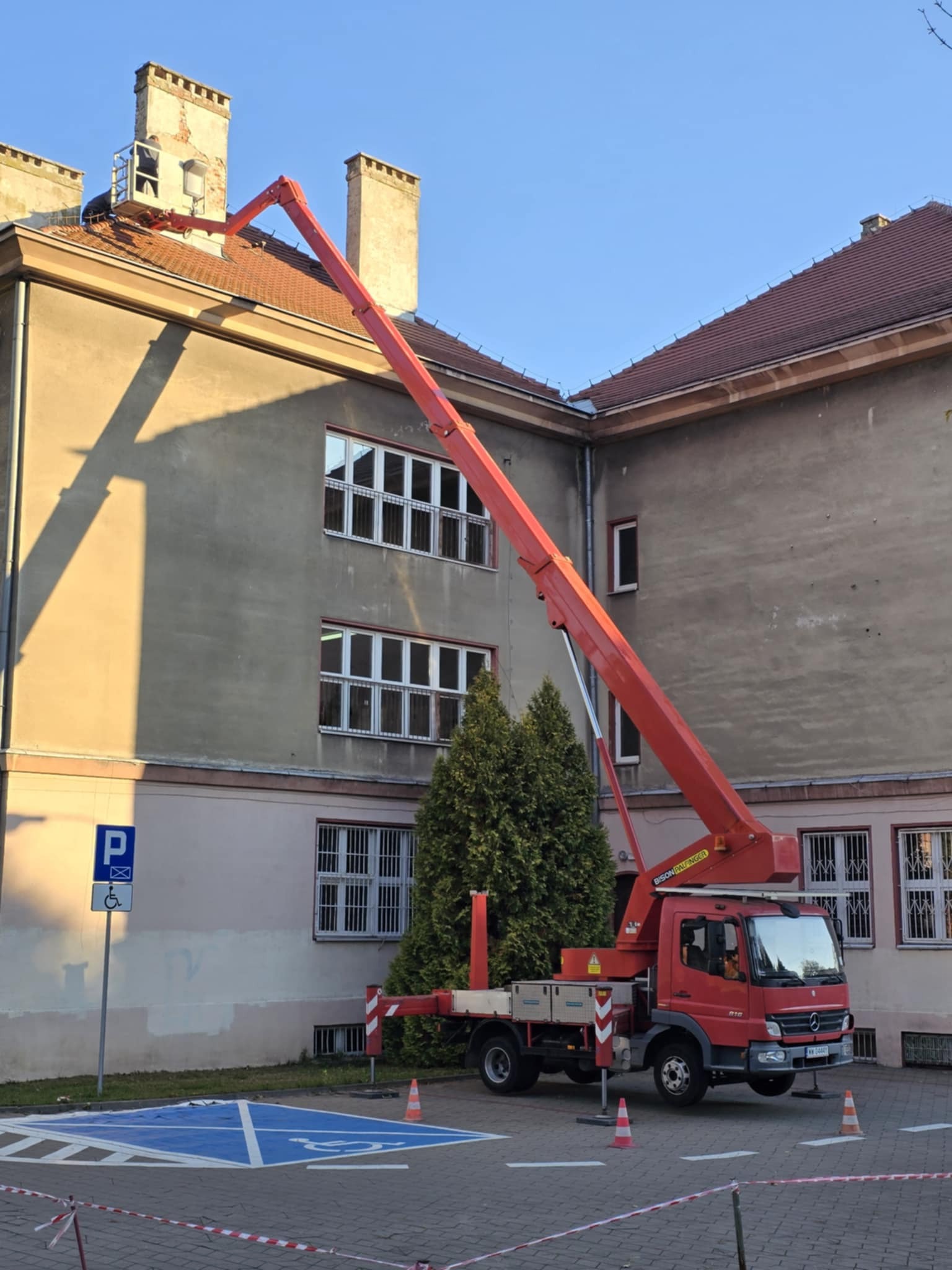 Czerwony pojazd z wysięgnikiem parkuje w Komorowie, przy budynku z ceglanym dachem i kominami, gdzie na platformie wysięgnika znajduje się osoba pracująca przy kominie.