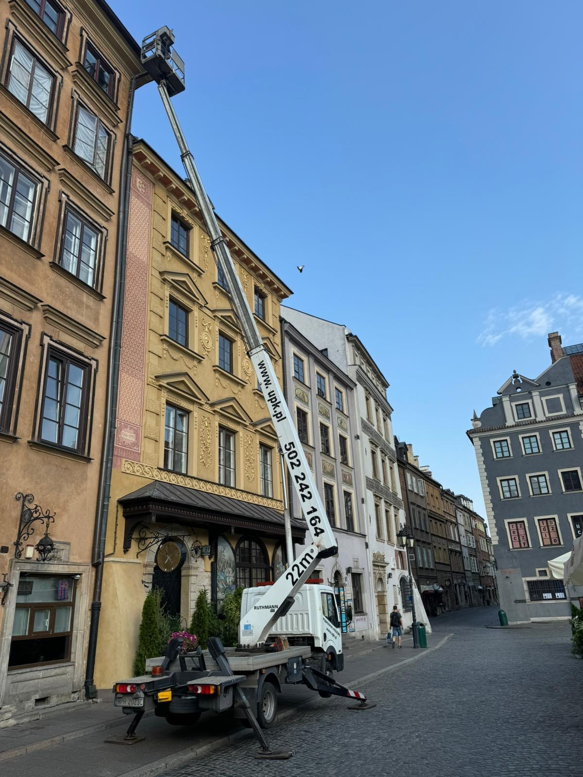 Podnośnik koszowy Ruthmann na tle kamienic Starego Miasta w Warszawie, z numerem telefonu i adresem strony internetowej na wysięgniku.