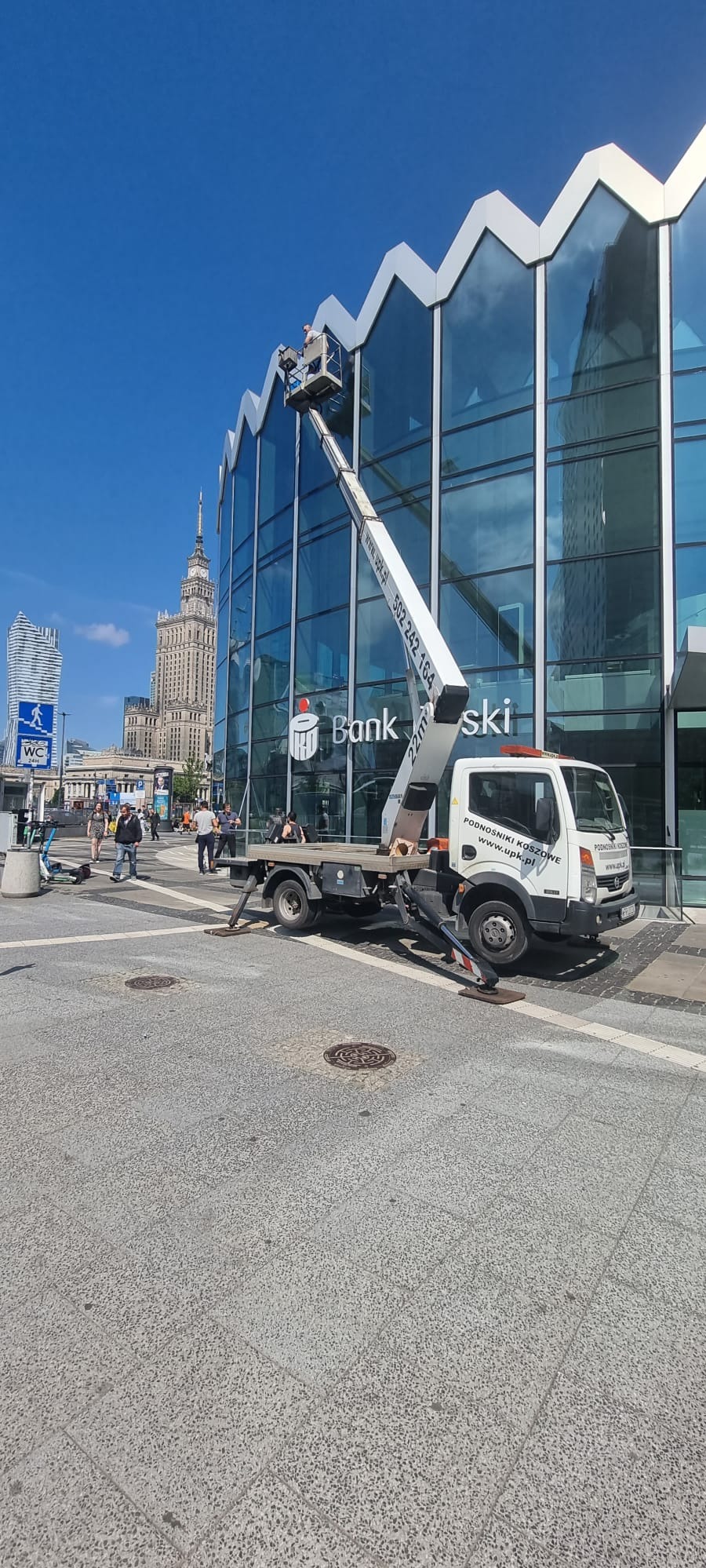 Pracownik w koszu na wysięgniku przy szklanej fasadzie budynku banku w centrum miasta, w tle Pałac Kultury i Nauki.