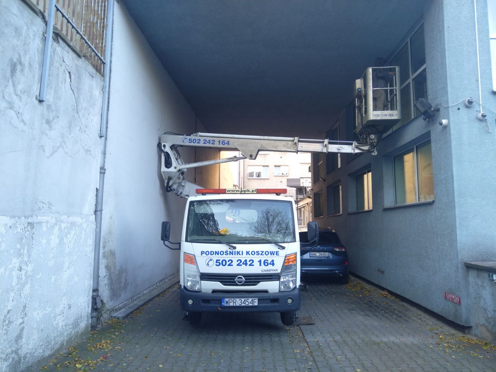 Biały samochód z wysięgnikiem parkuje w wąskim przejeździe między budynkami, platforma z pracownikiem unosi się przy ścianie budynku z oknami.