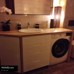 Łazienka z blatem drewnianym, umywalką nablatową, szafką z szufladami w kolorze kremowym i pralką w zabudowie, oświetlona dekoracyjną lampą.