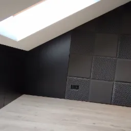 Pokój z panelami akustycznymi w odcieniach szarości i czerni, oświetlony przez okno dachowe, prezentujący minimalistyczny design z ekspozycją modeli samochodów na półce.