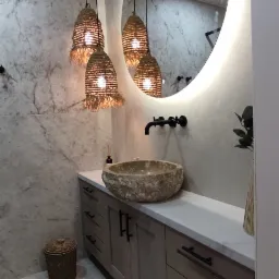 Łazienka z kamienną umywalką w kształcie misy, podświetlanym okrągłym lustrem i trzema wiszącymi lampami z abażurami z naturalnego materiału, szafka z szufladami i marmurowe ściany.