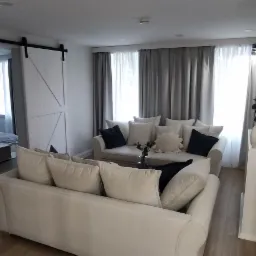 Jasne wnętrze salonu z dwoma białymi sofami, szarą zasłoną i przesuwnymi drzwiami do sypialni w tle.