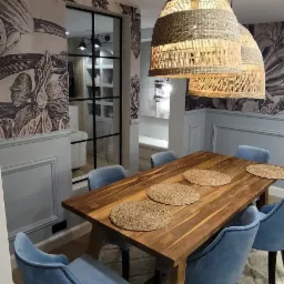 Jadalnia w stylu boho z drewnianym stołem, błękitnymi krzesłami obitymi aksamitem, plecionymi podkładkami i lampami z trawy morskiej. W tle ściana z tapetą w motywy roślinne i przeszklone drzwi.