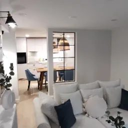 Jasne wnętrze domu z widokiem na kuchnię przez okno z czarną ramą, sofa z poduszkami w odcieniach bieli i granatu, drewniana podłoga i nowoczesne oświetlenie.