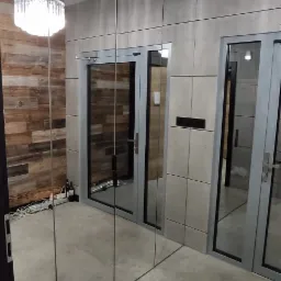 Nowoczesny korytarz z lustrzaną ścianą, dwoma drzwiami z szarymi ramami i drewnianą ścianą w tle, oświetlony dwoma kryształowymi lampami sufitowymi.