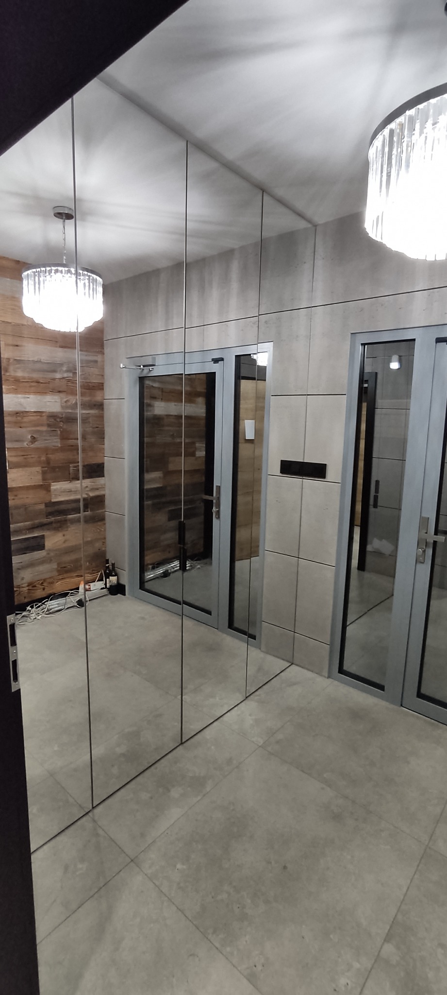 Nowoczesny korytarz z lustrzaną ścianą, dwoma drzwiami z szarymi ramami i drewnianą ścianą w tle, oświetlony dwoma kryształowymi lampami sufitowymi.
