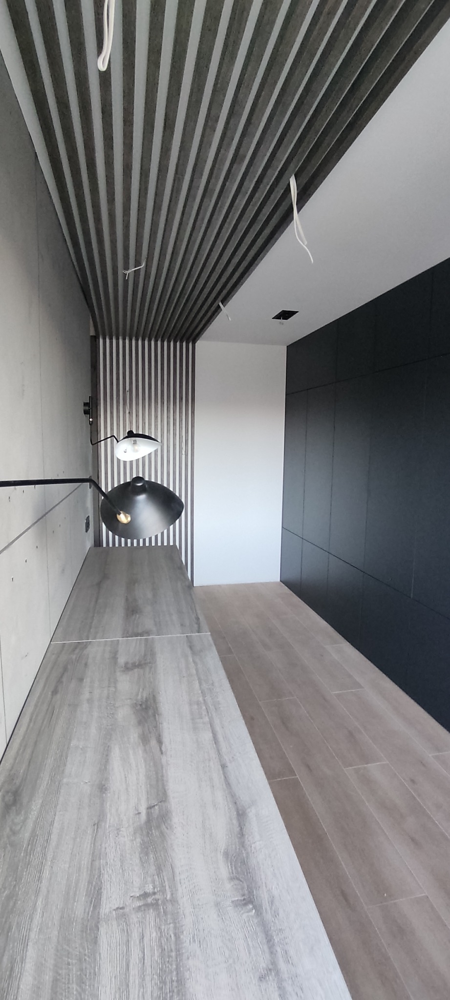 Nowoczesne wnętrze z drewnianym sufitem lamelowym, długim blatem z szarego drewna, czarną lampą i zabudową ścienną w ciemnym kolorze, uzupełnione panelami podłogowymi w odcieniach beżu.