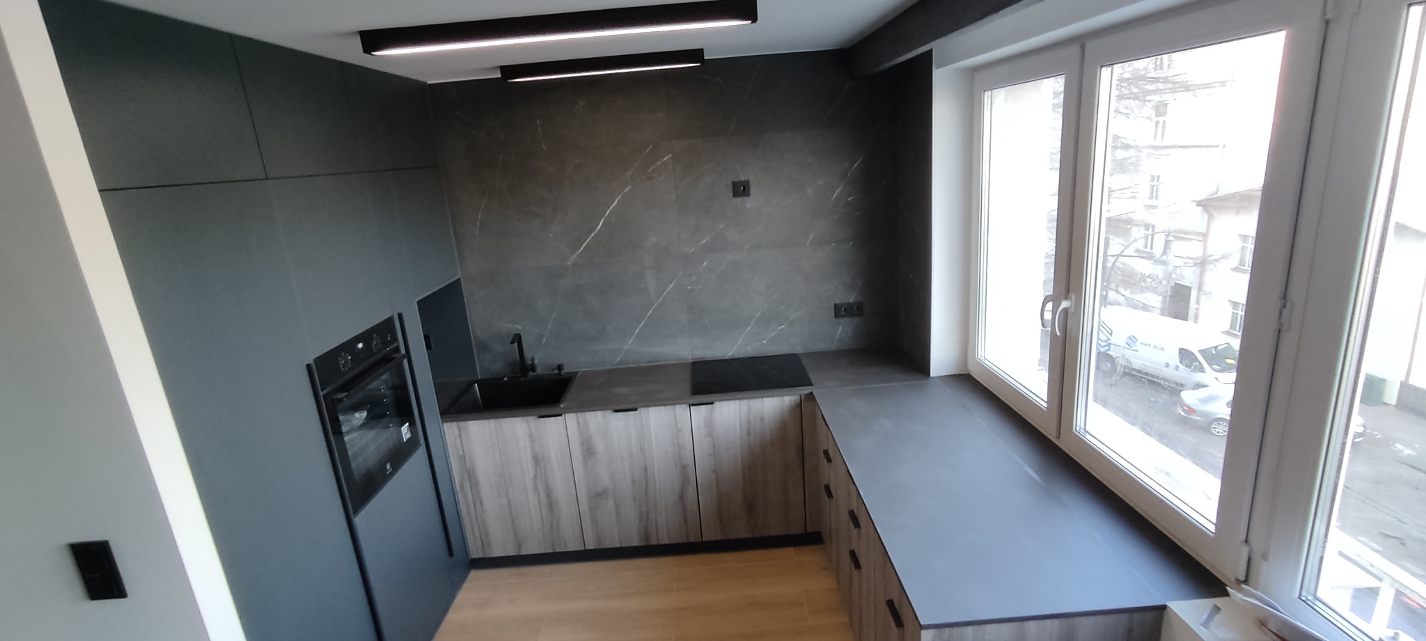 Nowoczesna kuchnia z szarymi frontami, drewnopodobnymi szafkami dolnymi i blatem zintegrowanym z parapetem okiennym. Widoczny czarny zlewozmywak, płyta indukcyjna i piekarnik w zabudowie.