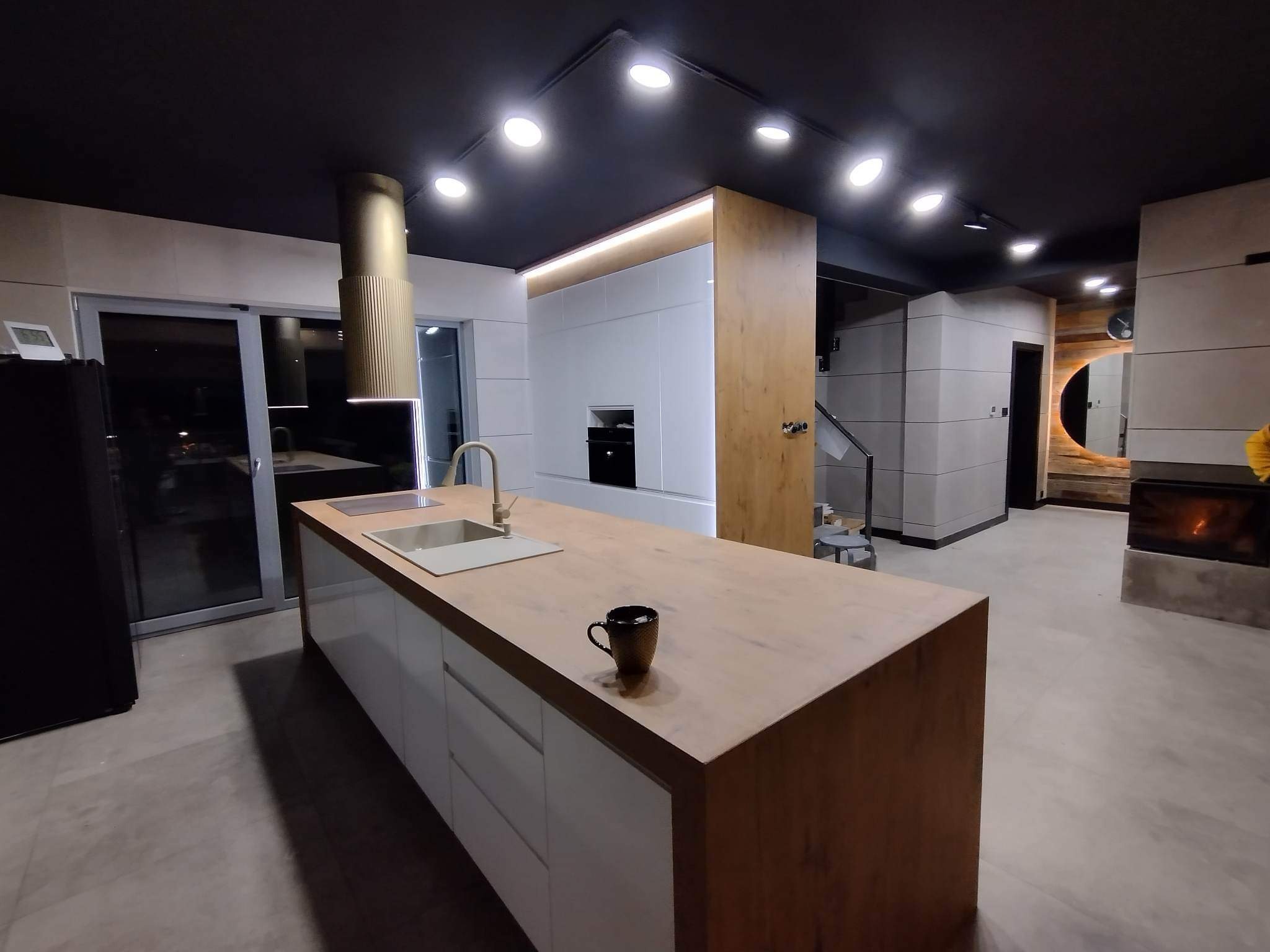Nowoczesna kuchnia z wyspą, wykończona w bieli i drewnie, z czarnym sufitem i oświetleniem punktowym. Na wyspie stoi czarny kubek, a w tle widoczny kominek i schody.