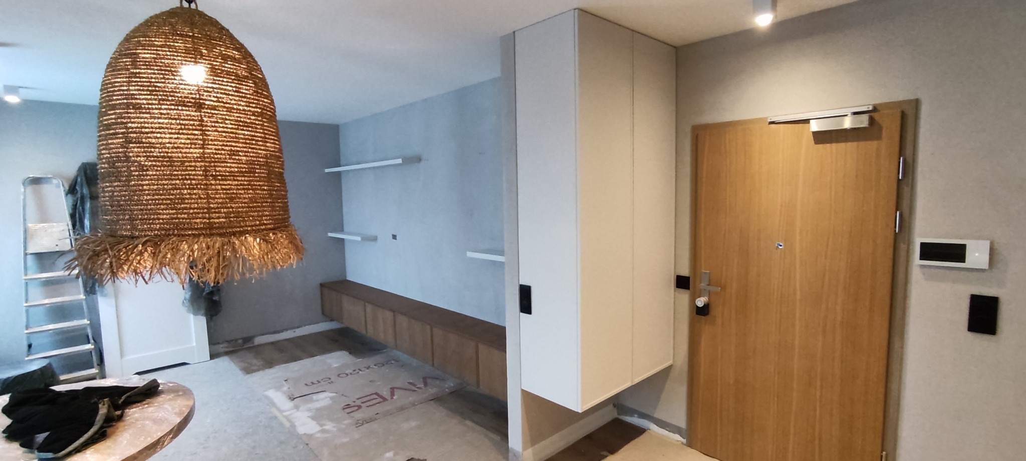 Wnętrze w trakcie wykańczania: wisząca szafka, drewniana szafka RTV, półki na ścianie, drewniane drzwi wejściowe, lampa z rafii, w tle drabina malarska i kartony z glazurą.
