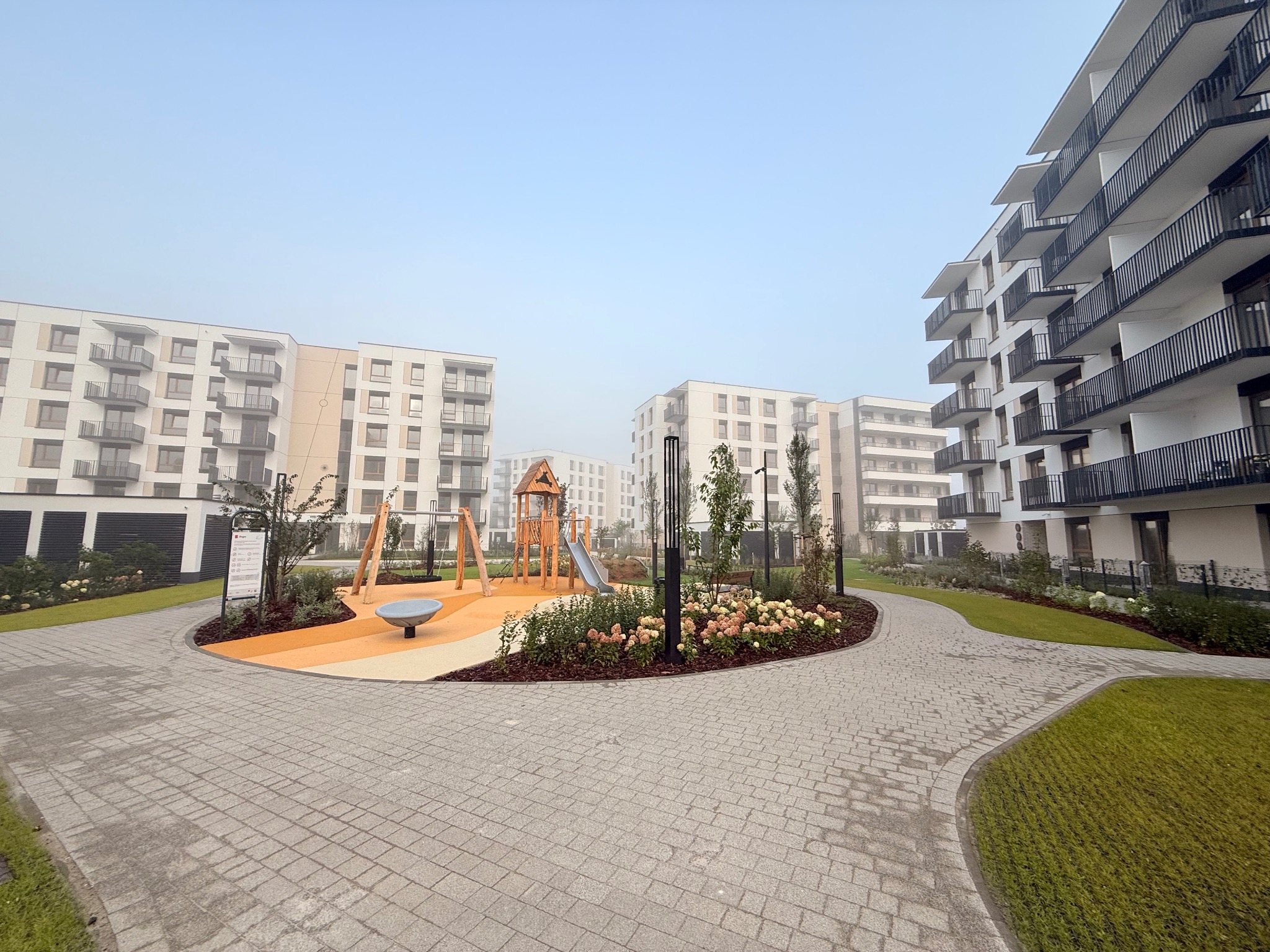 Nowoczesne osiedle w Pruszkowie z placem zabaw i ścieżką z kostki brukowej. Widok na budynki w lekkiej mgle, z dbałością o detale wykończenia.