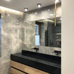 Nowoczesna łazienka z szarymi płytkami imitującymi beton, podwójną umywalką z czarnym blatem i drewnianymi szafkami, czarną armaturą i oświetleniem punktowym.