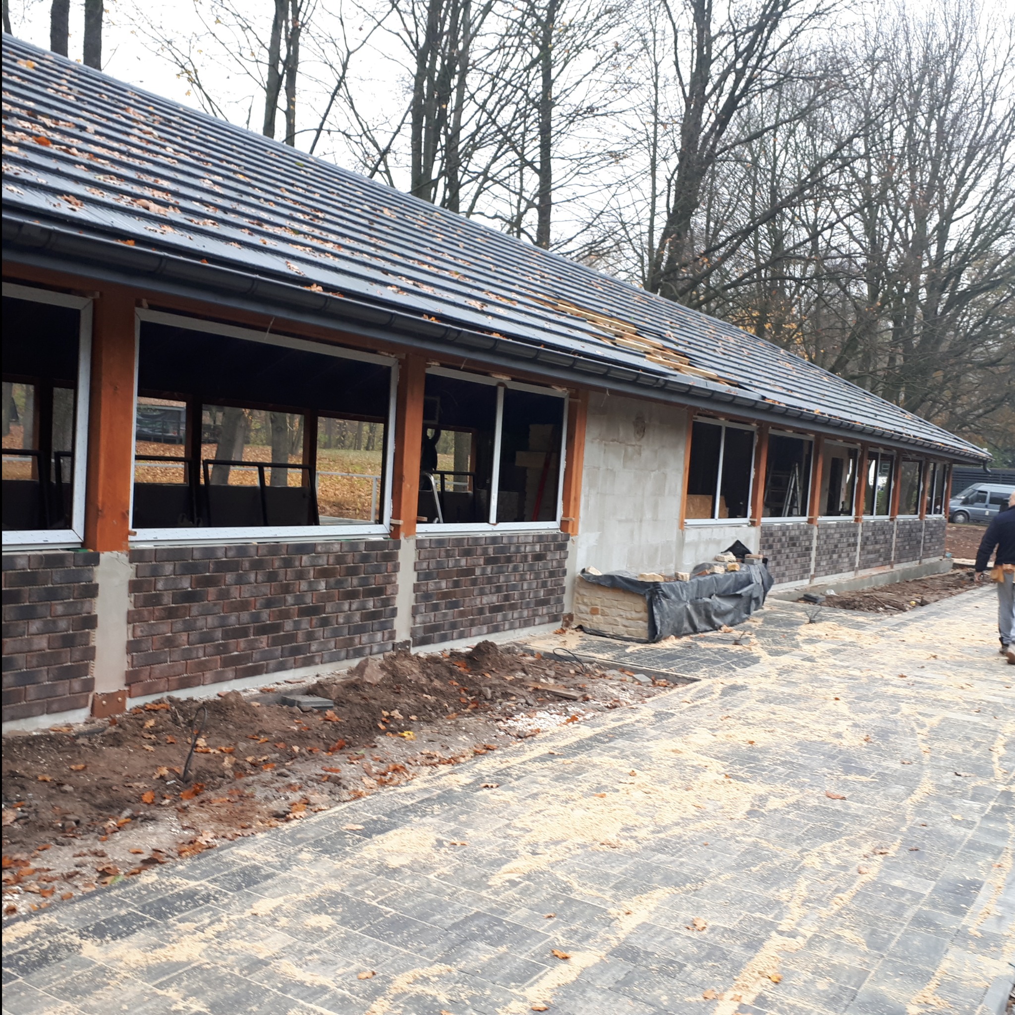 Budynek w trakcie wykończenia elewacji z cegły klinkierowej, widoczne okna w białych ramach, częściowo otynkowana ściana i ułożona kostka brukowa przed budynkiem.