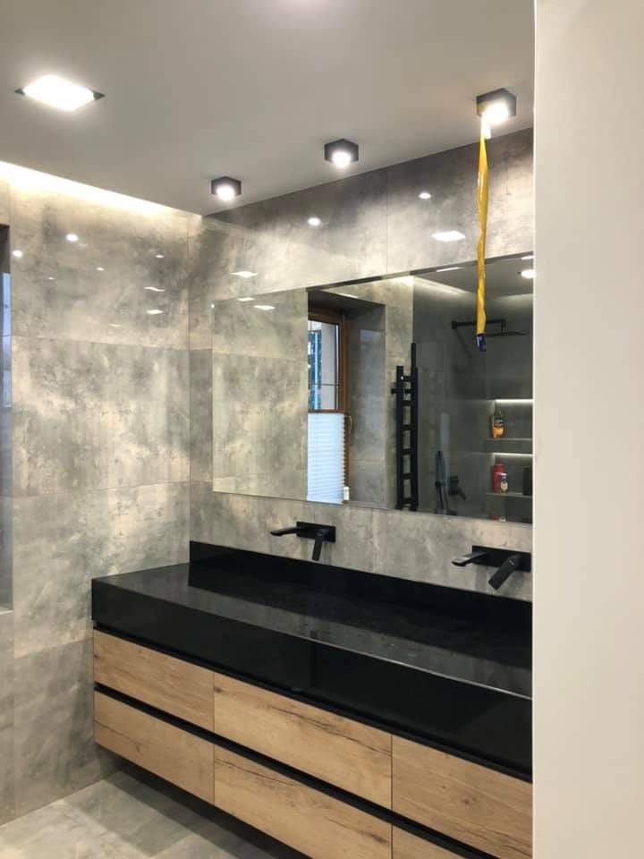 Nowoczesna łazienka z szarymi płytkami imitującymi beton, podwójną umywalką z czarnym blatem i drewnianymi szafkami, czarną armaturą i oświetleniem punktowym.