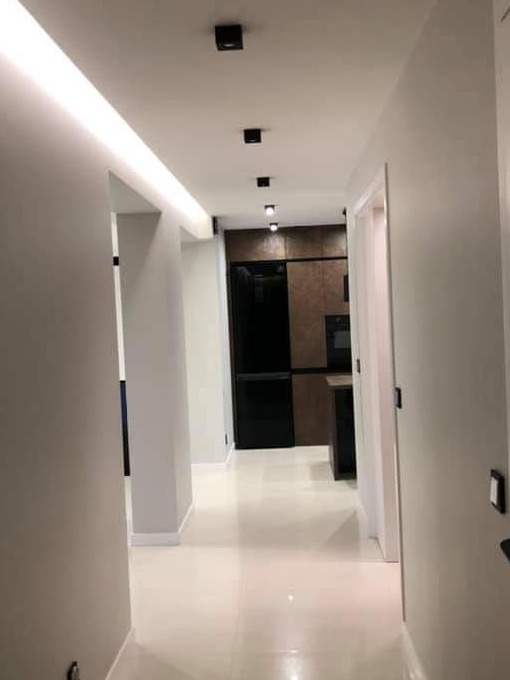 Nowoczesny korytarz z białymi ścianami i błyszczącą podłogą prowadzący do kuchni z czarną lodówką i brązowymi szafkami, oświetlony punktowymi lampami sufitowymi.