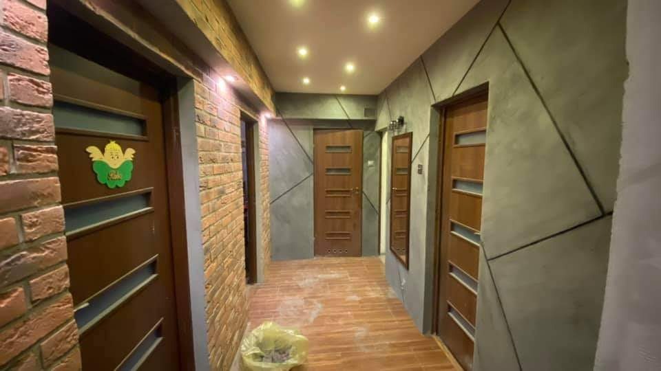 Korytarz po remoncie z ceglaną ścianą, szarymi ścianami z geometrycznym wzorem, drewnianymi drzwiami z przeszkleniami i brązowymi płytkami na podłodze. Na jednych z drzwi widoczna dziecięca naklejka.