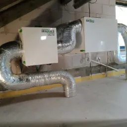 Dwa białe rekuperatory z logiem WANAS, połączone srebrnymi, elastycznymi przewodami wentylacyjnymi, zamontowane w surowym pomieszczeniu z oknem i odkrytą konstrukcją dachu.