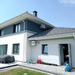 Nowoczesny dom dwukondygnacyjny z zamontowanymi jednostkami zewnętrznymi klimatyzacji na ścianach, widok z ogrodu, błękitny kompresor na kostce brukowej, zabawki dziecięce obok