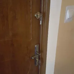 Brązowe skrzydło z zamontowanym srebrnym zamkiem i klamką, klucze w zamku, widoczny fragment ściany w neutralnym odcieniu i włącznik światła.