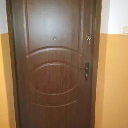 Ciemnobrązowe skrzydło z wizjerem i kluczem w zamku, osadzone w ościeżnicy, na tle ściany pomalowanej w pasy: biały i żółty.