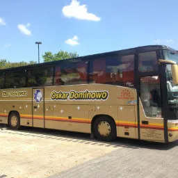 Autokar turystyczny w kolorze beżowym z napisem 'Biuro Turystyczne Oskar Dominowo' i logiem globusa na tle błękitnego nieba, zaparkowany na brukowanym placu.