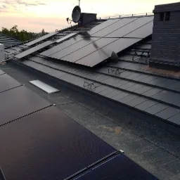 Instalacja paneli słonecznych na dachu pokrytym grafitową dachówką, widoczne elementy montażowe i komin z cegły, na tle zachodzącego słońca.