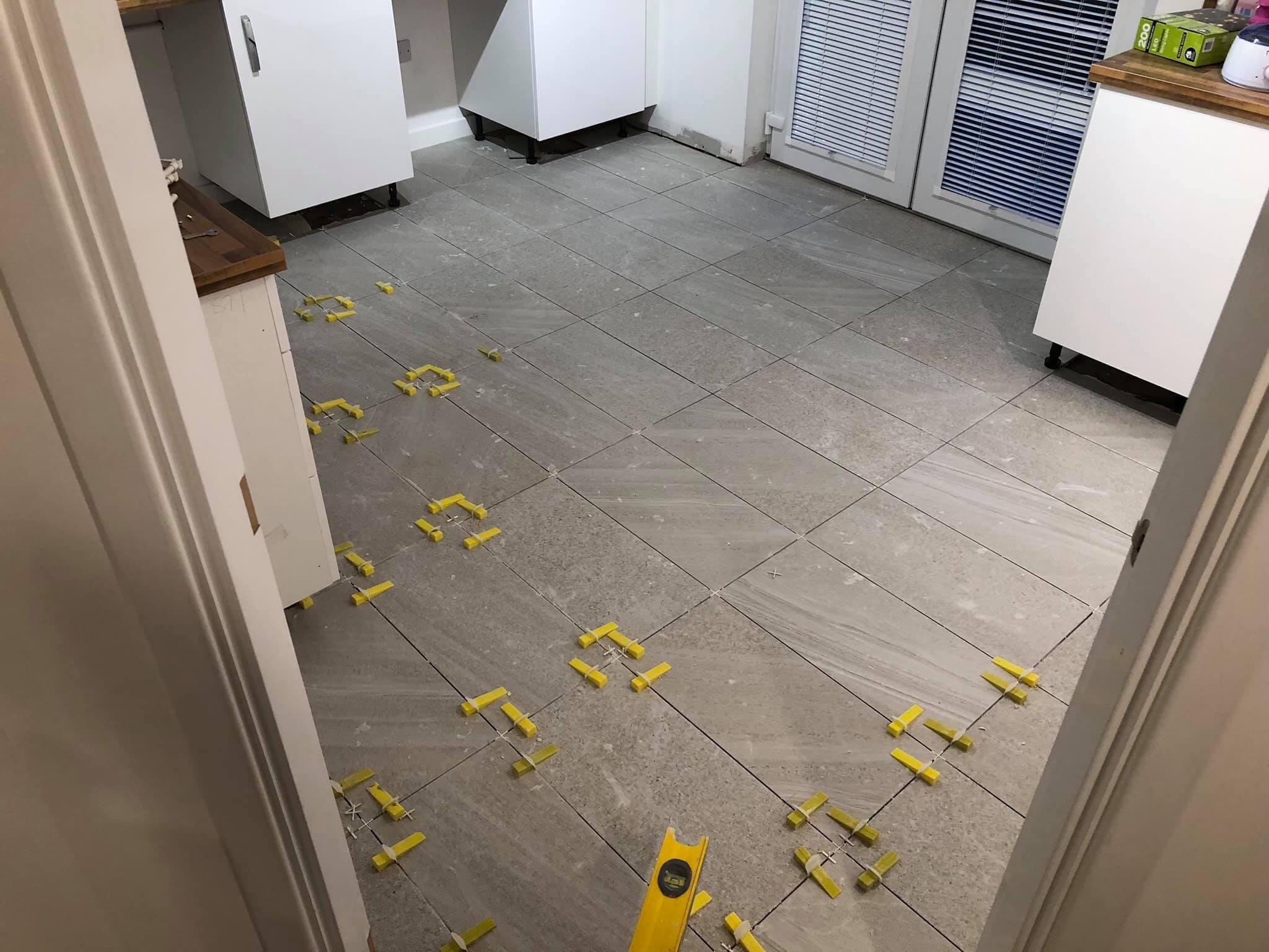 Układanie szarych płytek podłogowych w kuchni, widoczne białe szafki i żółte kliny dystansowe do płytek oraz poziomica