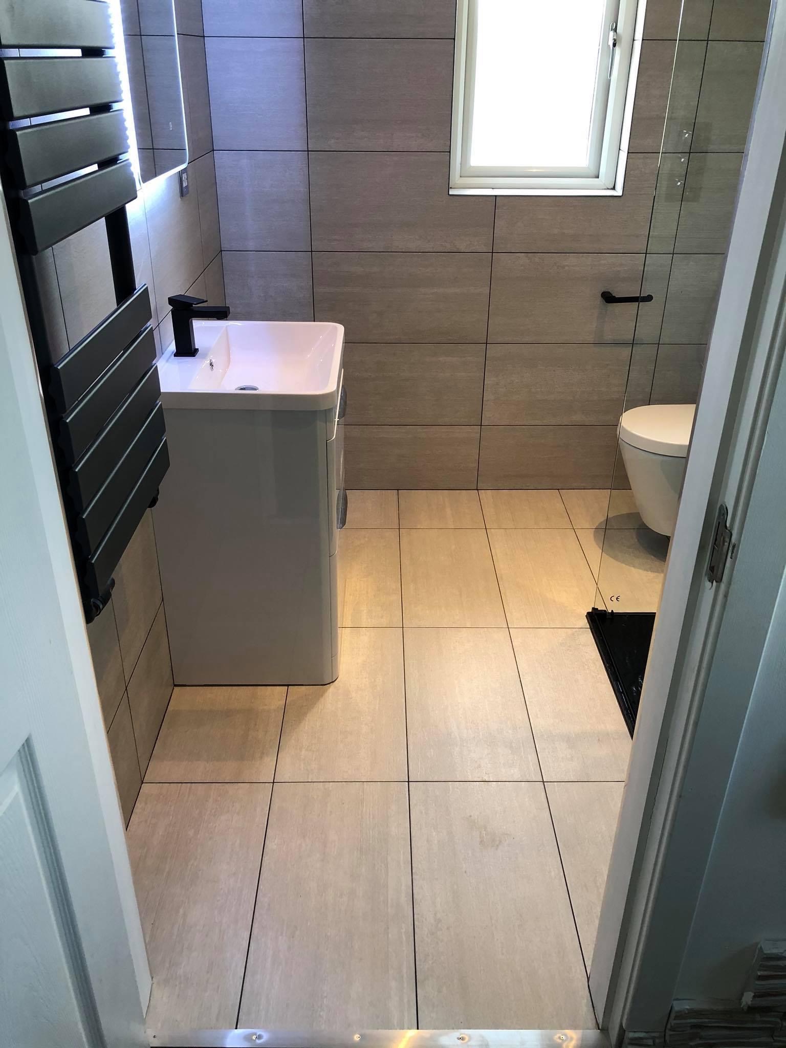 Nowoczesna łazienka z beżowymi płytkami imitującymi kamień, czarną armaturą i grzejnikiem drabinkowym, widok zza uchylonych drzwi.