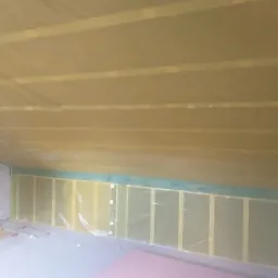 Poddasze w trakcie ocieplania, widoczne drewniane belki konstrukcyjne, izolacja pokryta folią, przewody elektryczne wystające ze ściany, fragment płyty gipsowej na podłodze.