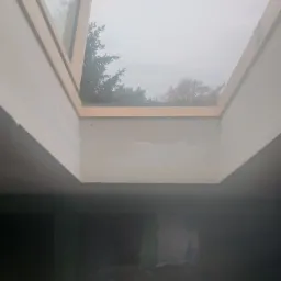 Wnętrze z oknem dachowym w drewnianej ramie, ściany przygotowane do malowania, widok na pochmurne niebo i drzewa.