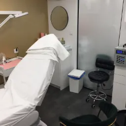 Kozetka kosmetyczna przykryta białym prześcieradłem w gabinecie z lampą powiększającą, okrągłym lustrem i oczyszczaczem powietrza.