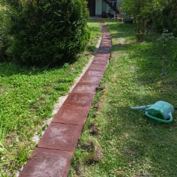 Ścieżka z brązowych płyt ułożona w ogrodzie, widoczna konewka i trawnik z koniczyną, dom w tle.