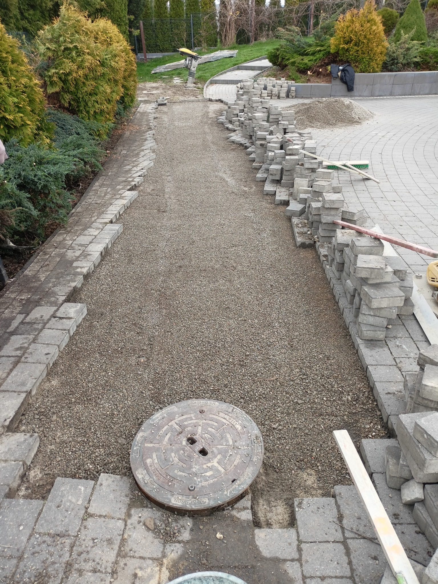 Etap budowy ścieżki z kostki brukowej: wyrównane podłoże z kruszywa, ułożone obrzeża, stosy kostki czekające na montaż, widoczna studzienka kanalizacyjna, w tle roślinność ogrodowa.