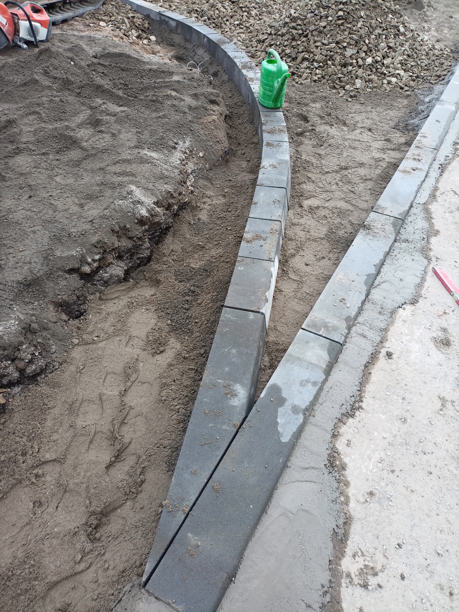 Wykop pod obrzeża z ułożonymi krawężnikami, zielony kanister na obrzeżu, przygotowane podłoże i piasek, widoczna piła do cięcia kamienia