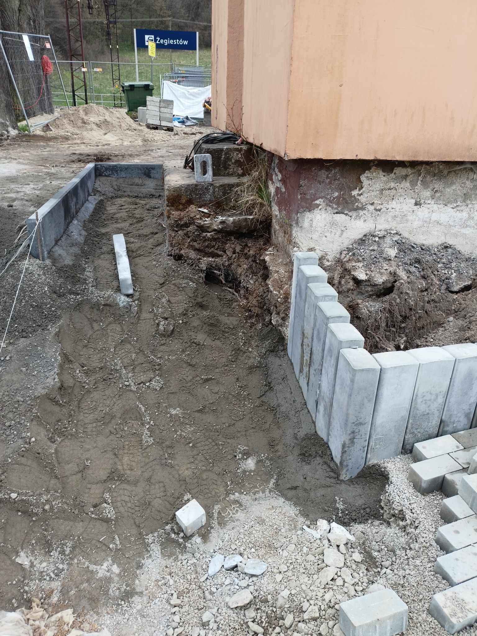 Przygotowanie podłoża pod ścieżkę z betonowych elementów obok budynku, widoczne obrzeża i częściowo ułożone elementy, w tle znak Zegiestów.
