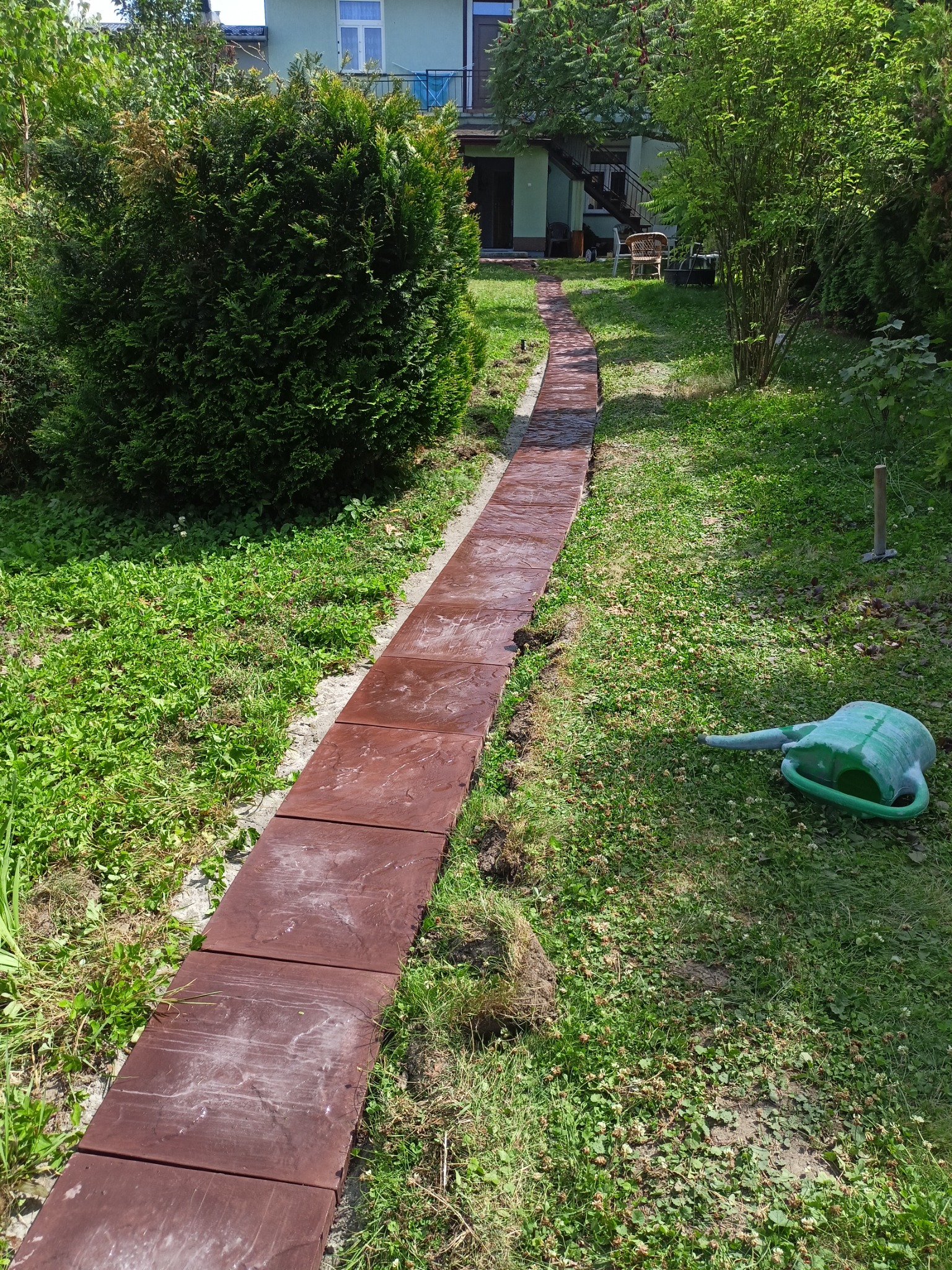 Ścieżka z brązowych płyt ułożona w ogrodzie, widoczna konewka i trawnik z koniczyną, dom w tle.
