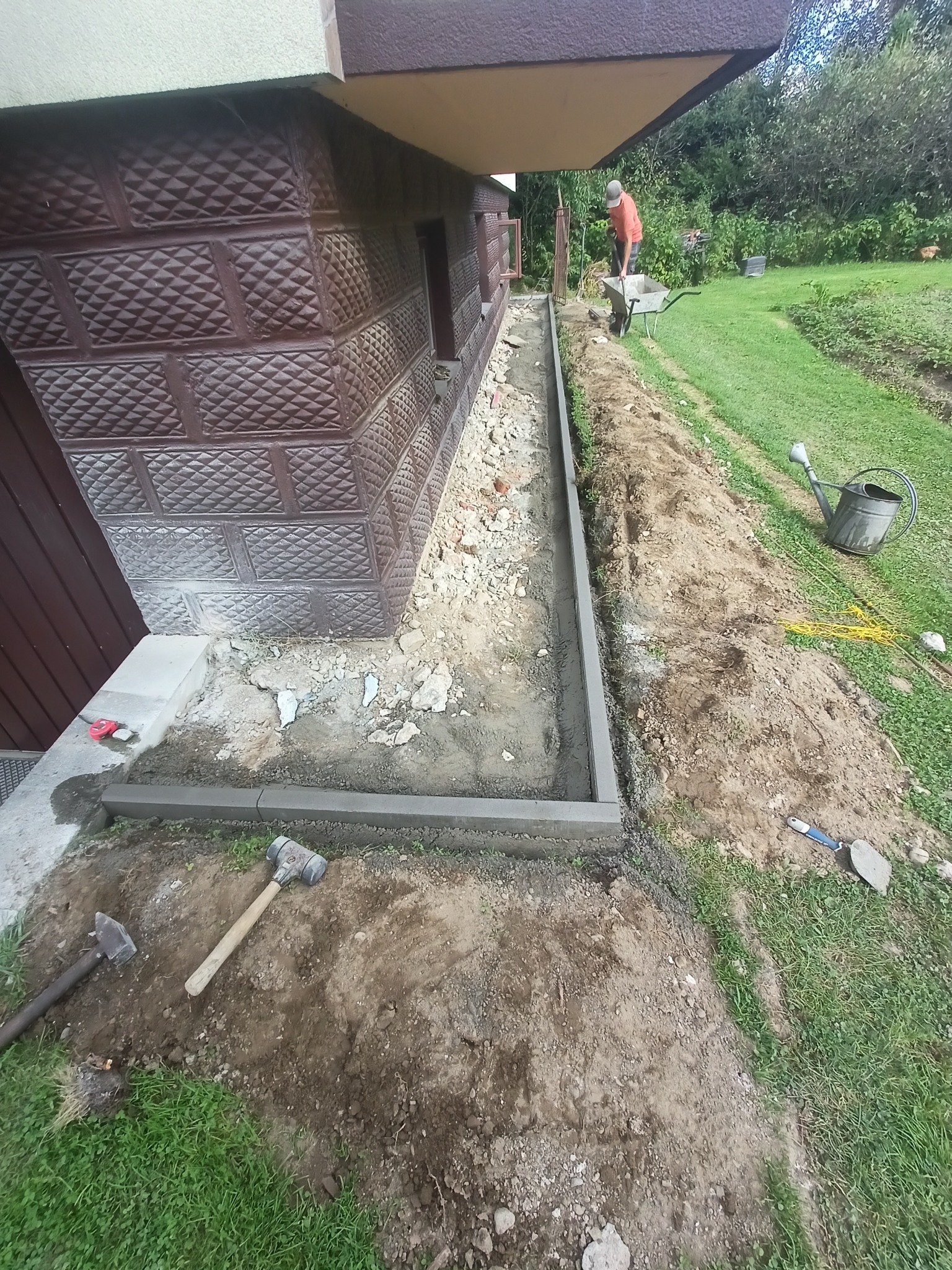 Przygotowanie podłoża pod obrzeża chodnika przy budynku z dekoracyjną, brązową elewacją; widoczny pracownik z taczką w tle, narzędzia budowlane leżące na trawie.