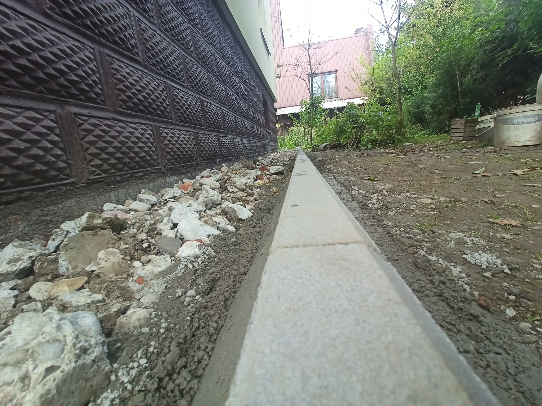 Świeżo ułożony betonowy obrzeże chodnika przy ścianie budynku z dekoracyjnym wykończeniem, widoczne gruz i ziemia po pracach ziemnych.