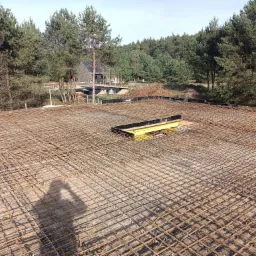 Siatka zbrojeniowa na betonowej płycie, widok z góry, w tle las i budynek w stylu nowoczesnej stodoły.