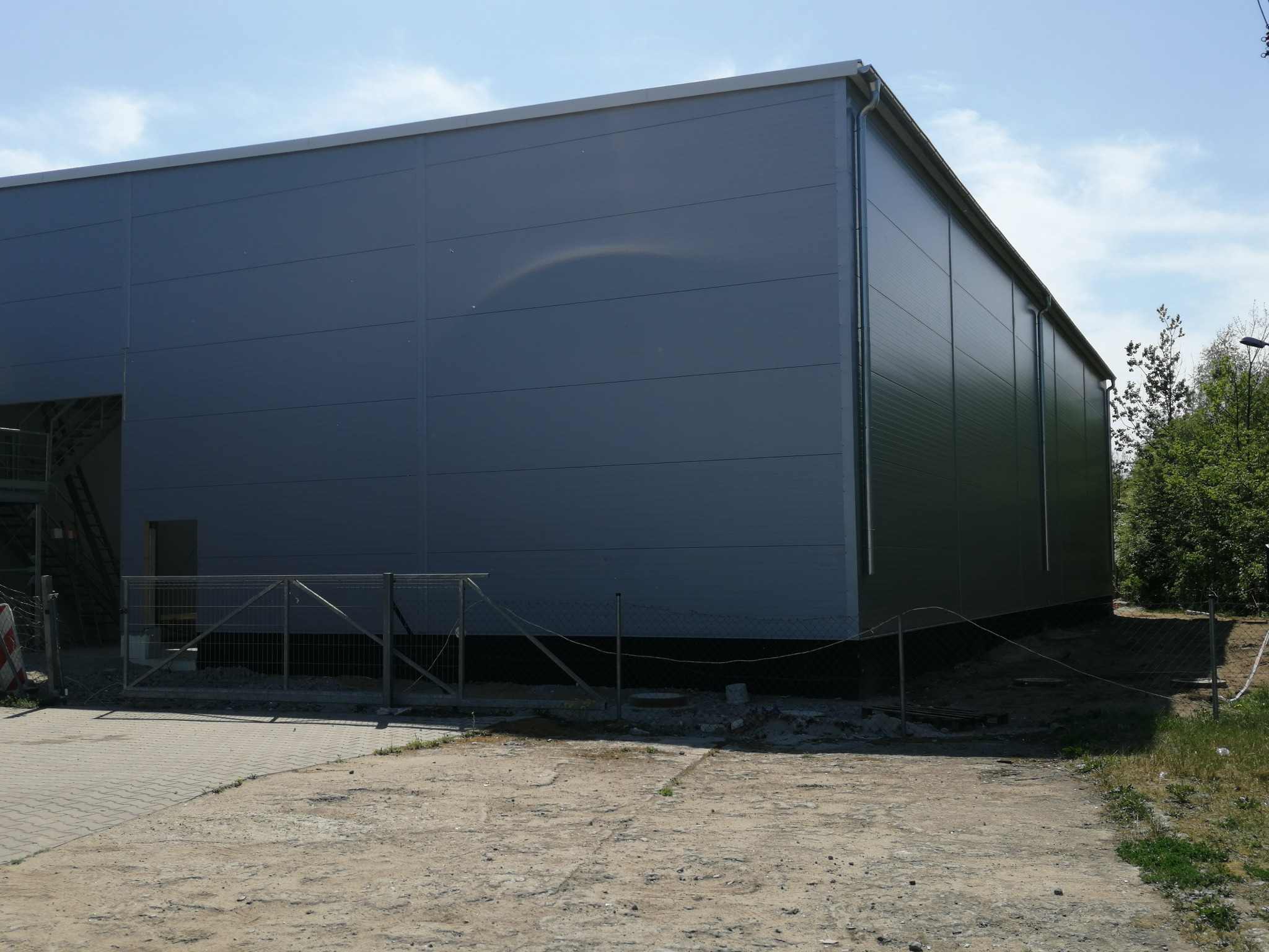 Nowoczesny budynek przemysłowy z paneli w kolorze szarym, widoczne łączenia paneli, z fragmentem ogrodzenia tymczasowego i terenem budowy w pierwszym planie.