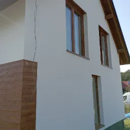 Świeżo otynkowana elewacja domu jednorodzinnego z drewnianymi ramami okiennymi i fragmentem drewnianej okładziny na rogu, widoczne zwisające kable elektryczne.